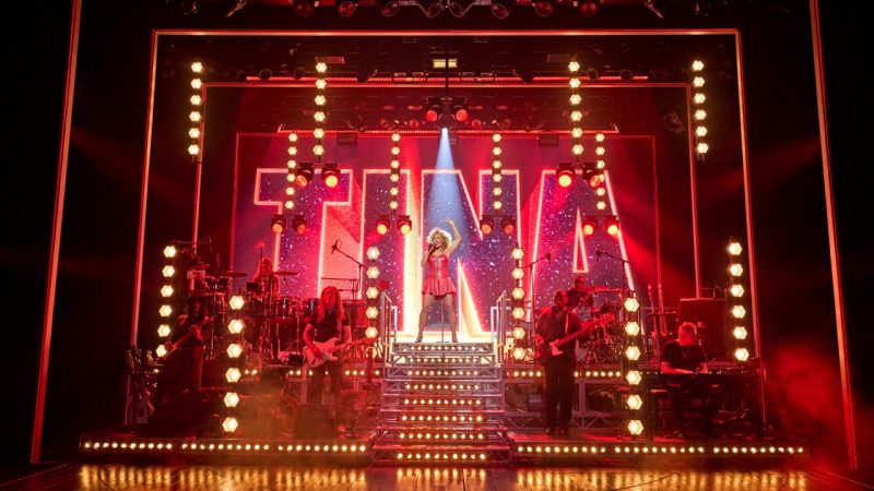 Cast members of "TINA – The Tina Turner Musical" perform on stage, giant lit letters reading TINA behind them.