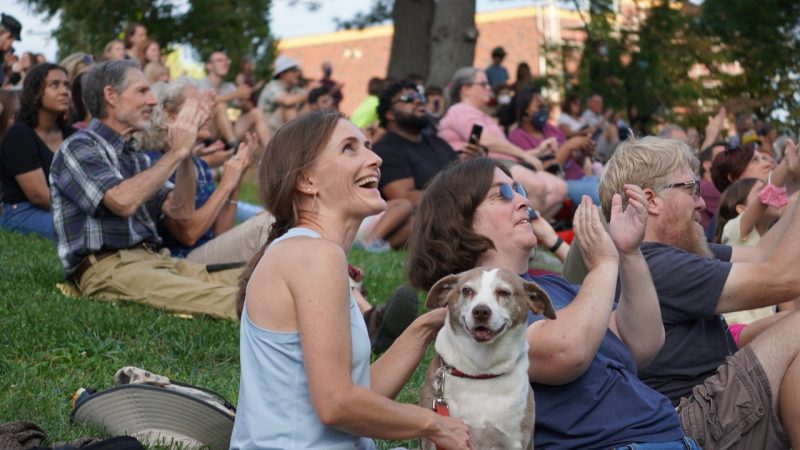  A crowd of people sits on the grass lawn of the Moss Arts Center, cheering and clapping. In the foreground, a white woman in a blue tank top holds her dog's leash and smiles widely, looking up towards where dancers performed on the side of the Moss Arts Center.