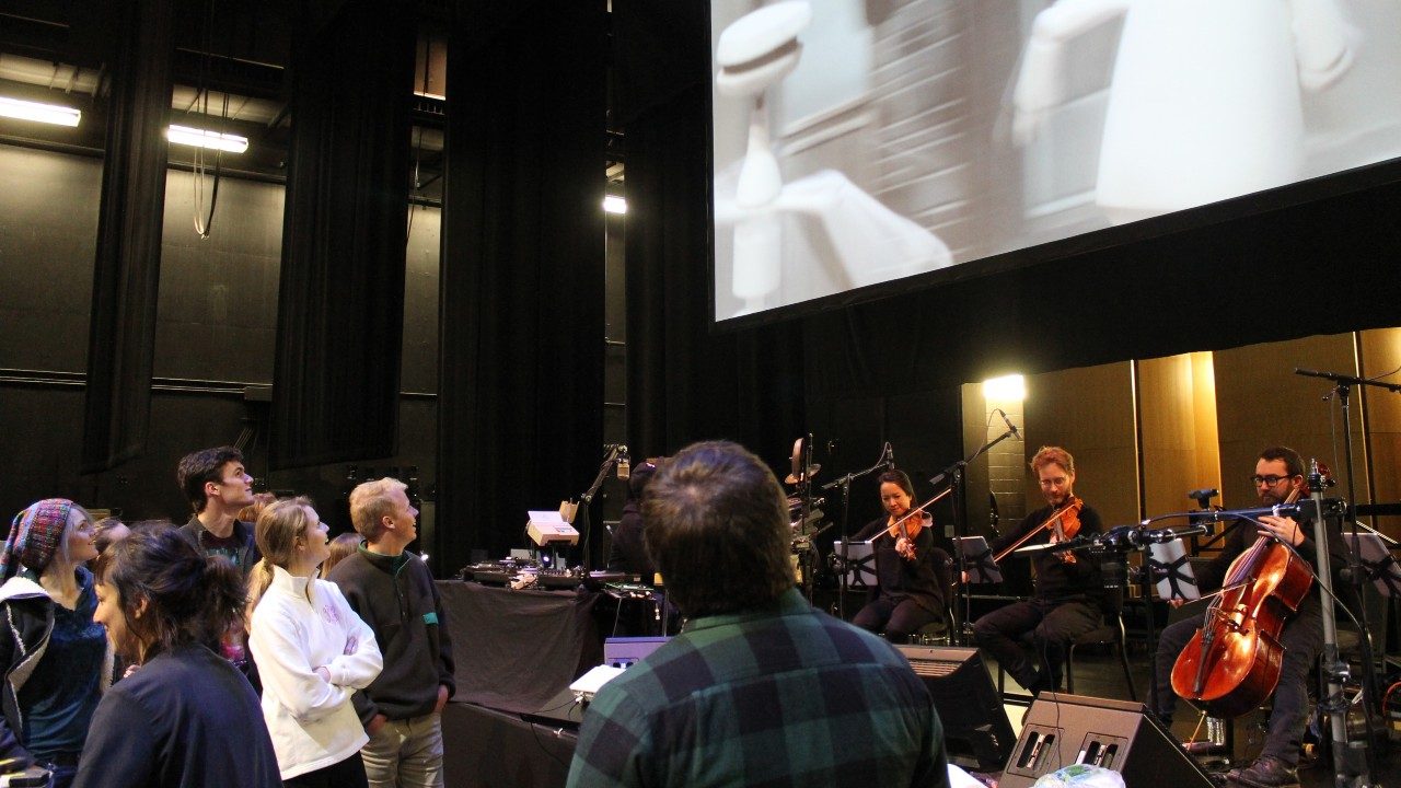  People standing in the Cube looking up at a projection of Kid Koala while musicians play underneath the projection screen.