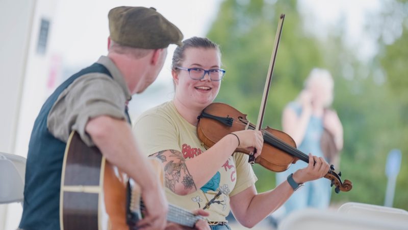 A young white woman playing a violin smiles and looks towards a white man in a vest and hat playing an acoustic guitar outside at the Moss Arts Center.