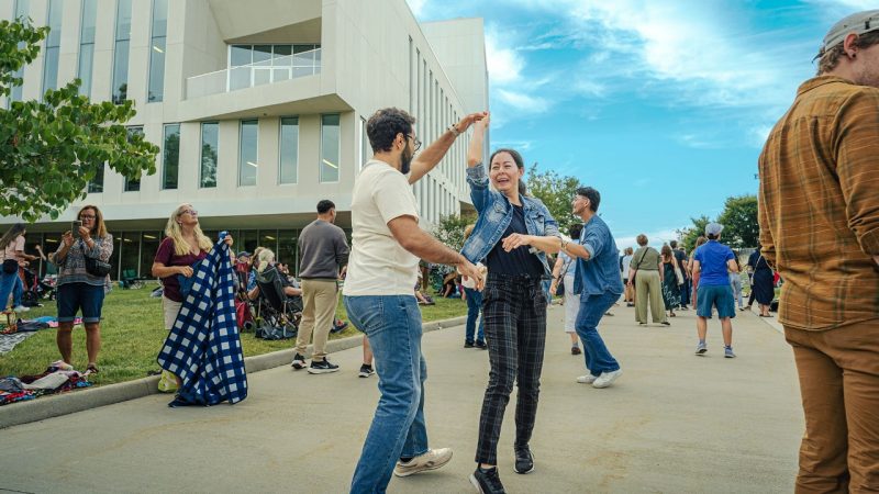 Community members, a man and a woman, dance during a free outdoor concert by Puerto Rican band Plena Libre at the Moss Arts Center under a blue sky.