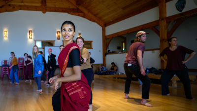 Members of Chitrasena Dance Company lead a dance workshop for students at Springhouse Community School in Floyd, Virginia. In the foreground, a brown woman with long dark hair wears a red costume wrapped over a black cropped shirt and pants and teaches high school-aged students in a large room with a vaulted wood ceiling.