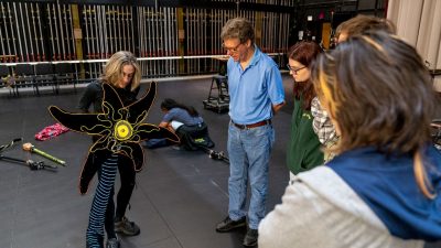 Virginia Tech students in a School of Performing Arts lighting design class meet with members of Lightwire Theater to learn tricks of the trade. A white woman with shoulder length blonde hair holds a light-up flower puppet and explains her creative process to three students and a professor.