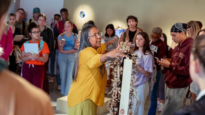 Virginia Tech students visit the Moss Arts Center galleries as a class. In the center, a middle aged Native woman in a yellow shirt discusses a sculptural piece on display.