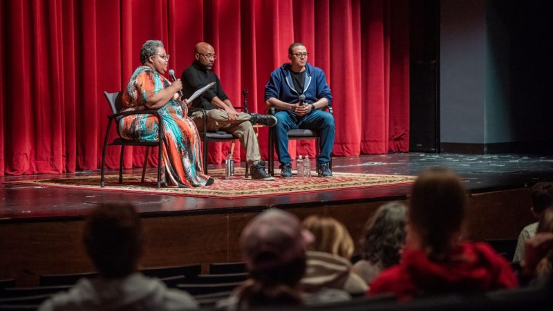 Members of Alvin Ailey American Dance Theater gave a talk before a performance at the Moss Arts Center in an event co-sponsored by the Black Cultural Center. Three people sit on stage in front of a red curtain while the audience looks on.