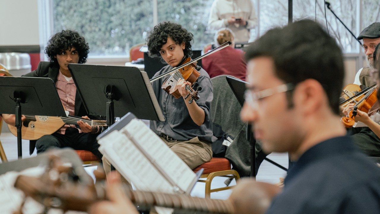  Members of Itraab Arabic Music Ensemble perform for Persian New Year. A brown man with short dark brown hair is out of focus in the foreground, and in the background two brown people with medium length curly dark brown hair play an oud, at left, and a violin, at right.