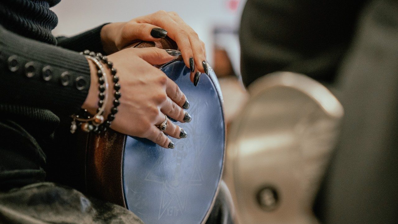 A close up of an Arabic lap drum played by a woman wearing a black sweater with long dark nails.