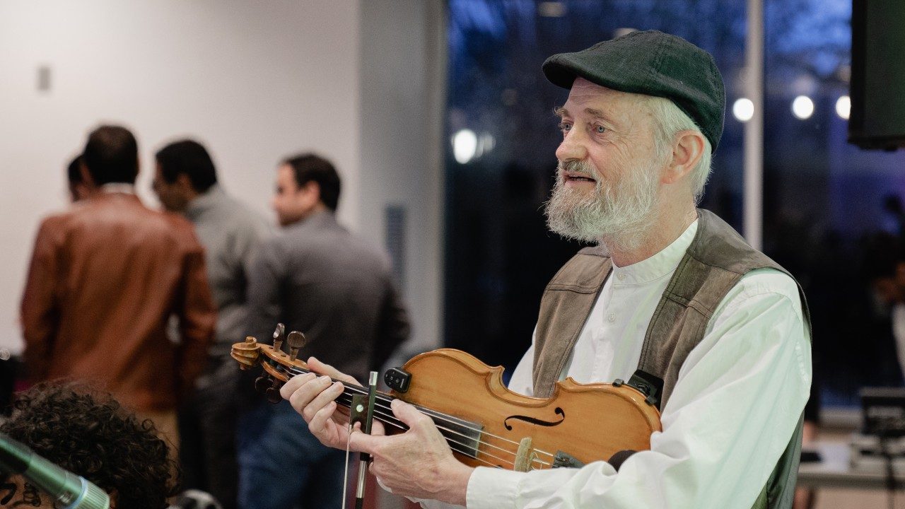  An older white man with white hair, a black hat, a white collarless button down shirt, and a light brown vest stands, holding a violin and bow.