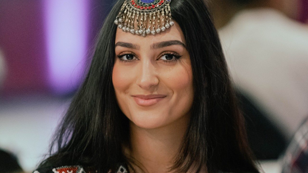A young brown woman with long, straight, dark brown hair and cat eyeliner wears a beaded headpiece and beaded traditional costume. She smiles towards the camera.