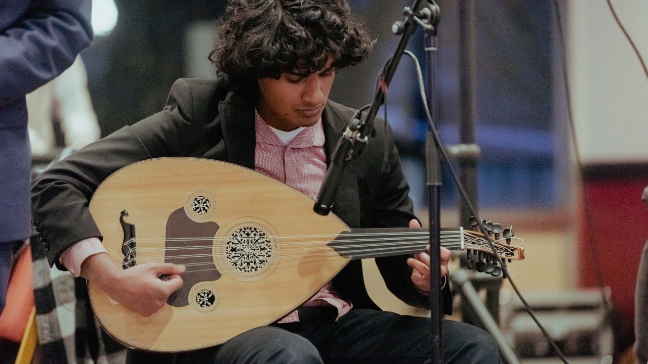  A young brown man with semi-long wavy brown hair plays an oud. He wears a dark brown blazer over a light pink button down shirt.