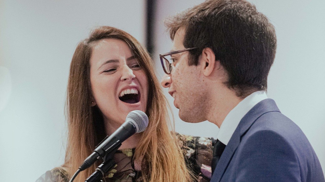  A woman, left, and a man, right, sing into a microphone together. The woman has long straight brown hair and sings fully. The man has short brown hair, glasses, and wears a navy coat over a white button down shirt.