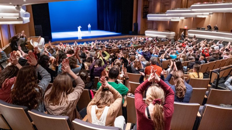 K12 students participate during a recent school-day performance, putting their palms together above their heads like shark fins at the instruction of two dancers on stage.