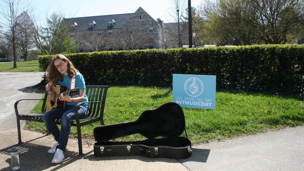  VT student plays guitar on campus during VT Music Day. Open guitar case sits next to her.