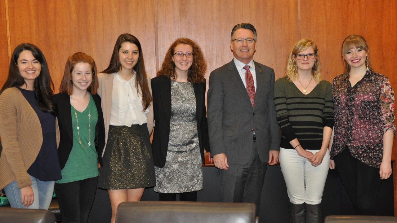  President Tim Sands posing with six Moss Arts Center student ambassadors in front of wood paneled wall.