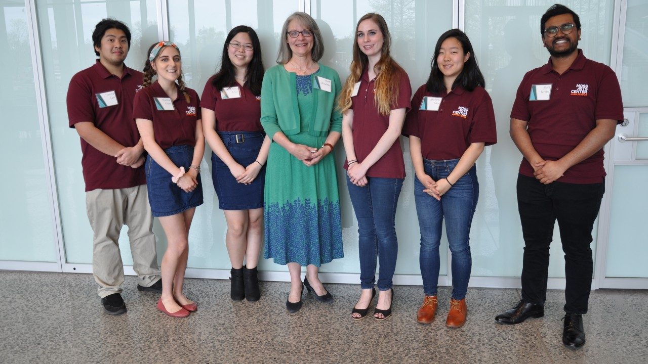 Six Moss Arts Center student ambassadors in their maroon shifts standing with Ruth Waalkes in the grand lobby.