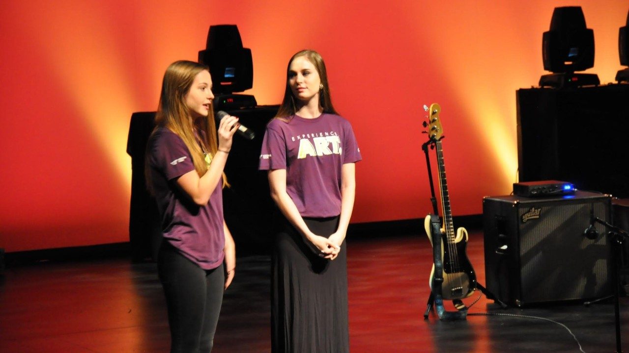  Two Moss Arts Center student ambassadors stand on the Fife stage giving a curtain speech. Stage is dark with a bright orange rear projection.
