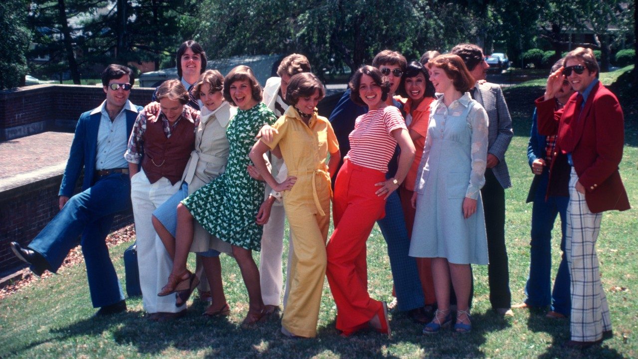 Members of the New Virginians gather in a group on a grassy clearing in the sun, dressed in peak 1970s attire.