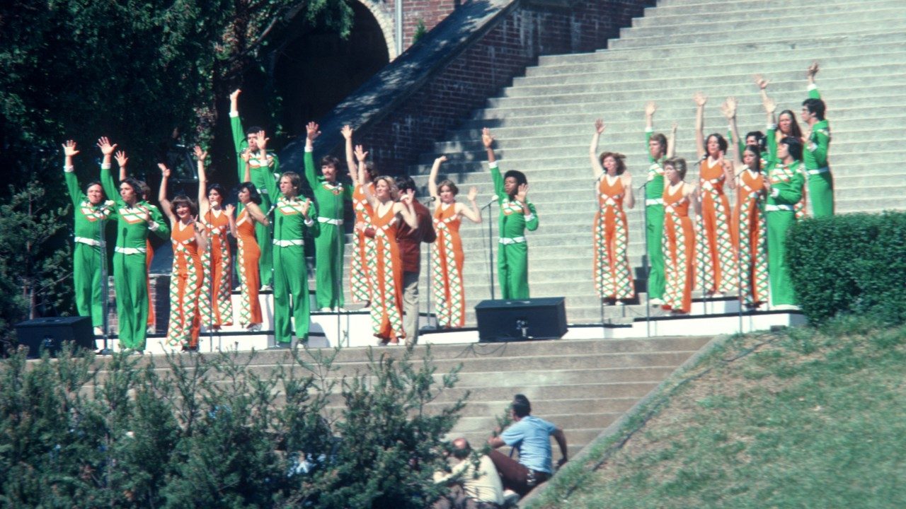  The New Virginians perform on the landing of a set of outdoor stairs. The men wear green coordinating sets, and the women wear orange and patterned coordinating sets. They stand at microphone stands with all have one arm raised in the air.