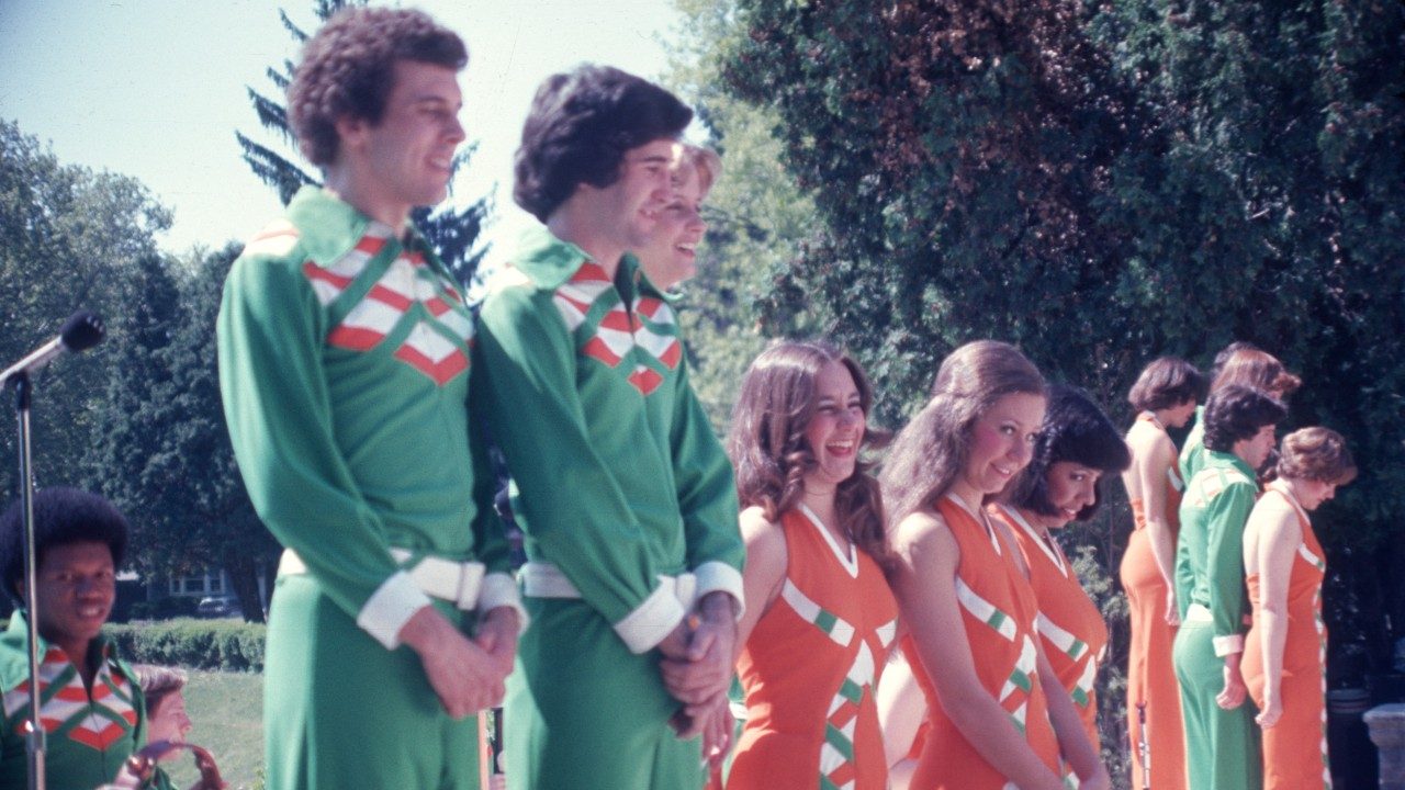  Some of the New Virginians perform or prepare to perform outdoors. Behind them are green, leafy trees. The men wear green pants and shirts; the women wear orange jumpers.