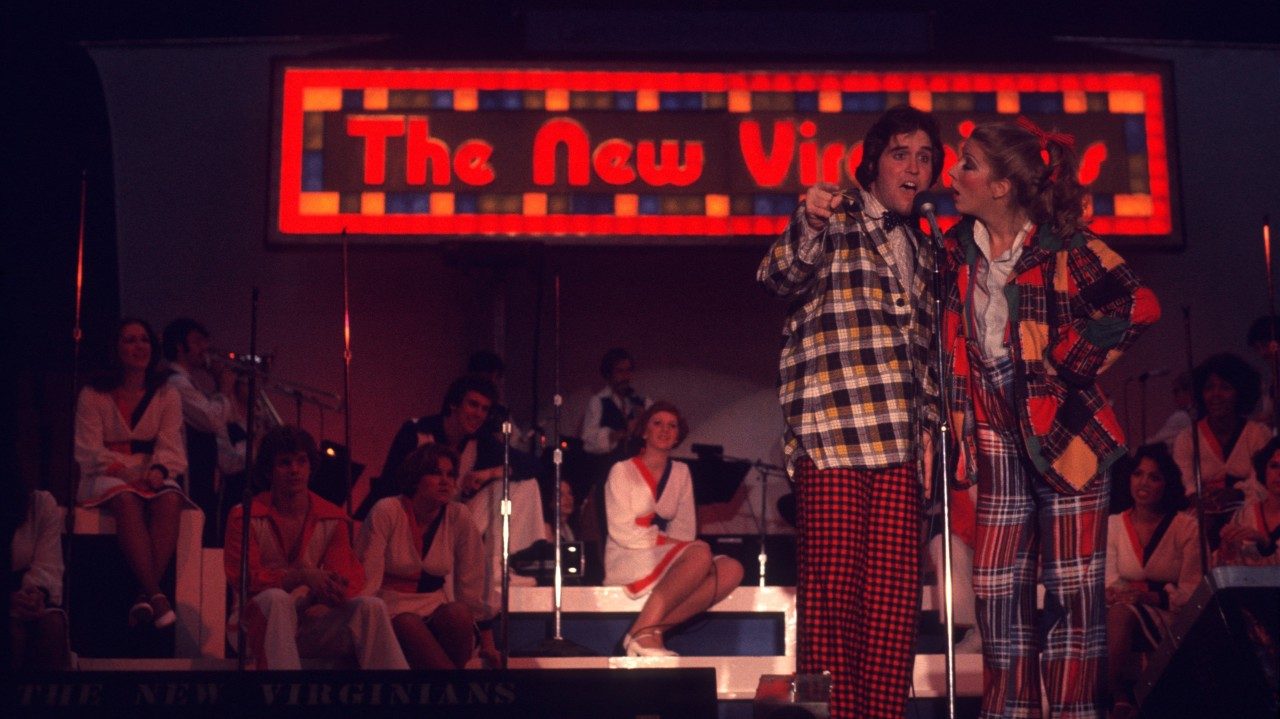  Members of the New Virginians perform on stage. Two people in the foreground sing into a microphone together: a white man with medium length dark brown hair, yellow and brown plaid shirt, and red and black plaid pants; and a women with blonde hair in a curled ponytail, multicolored plaid blazer, white button down shirt, and multicolored plaid pants. Behind them, other members sit on risers and a large light up sign reads "The New Virginians"