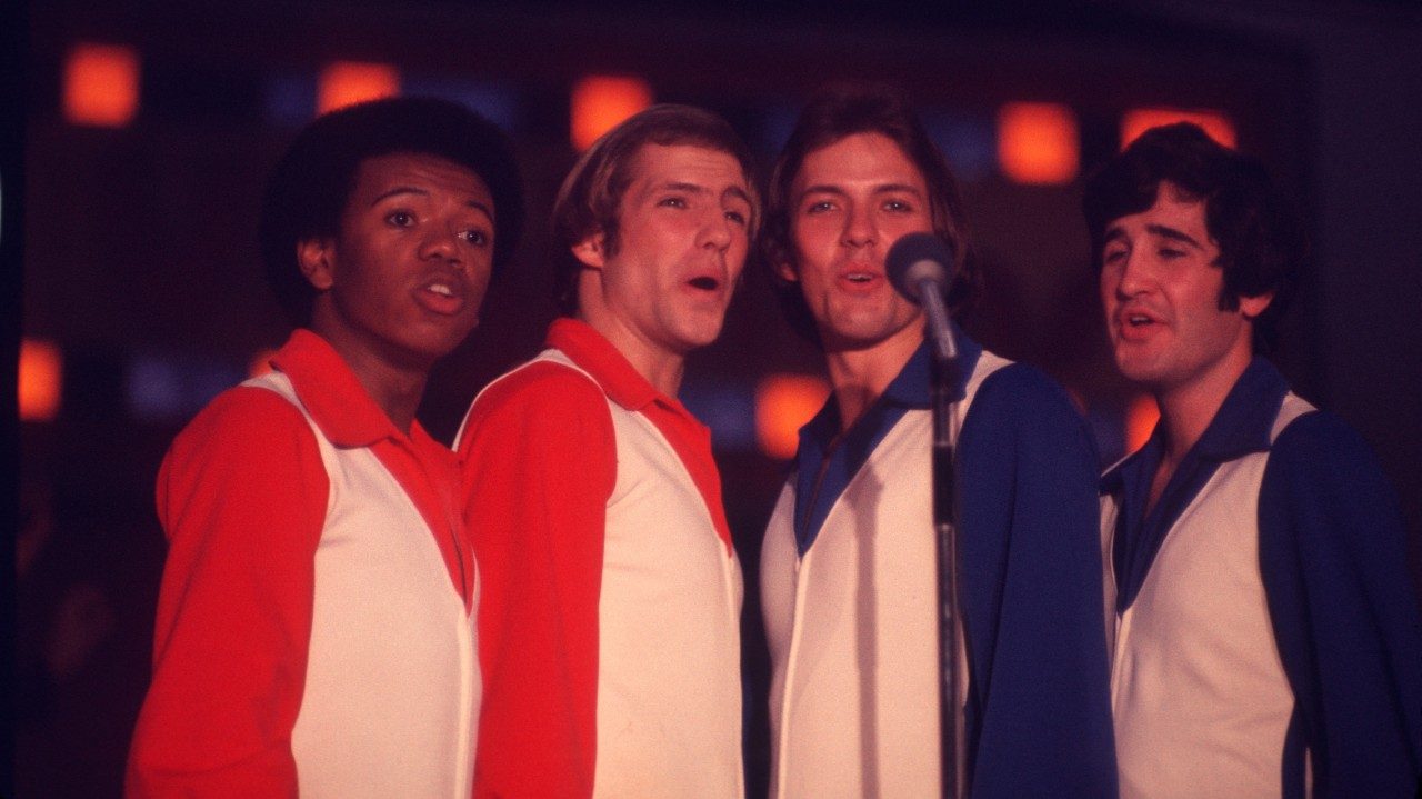  Four men from the New Virginians sing into the same microphone on stage. All four wear white vests over button down shirts. The two on the left, a Black man with a medium length afro and a white man with medium length blonde hair, wear red shirts. The two on the right, a white man with medium length light brown hair and a white man with medium length dark brown hair, wear blue shirts.