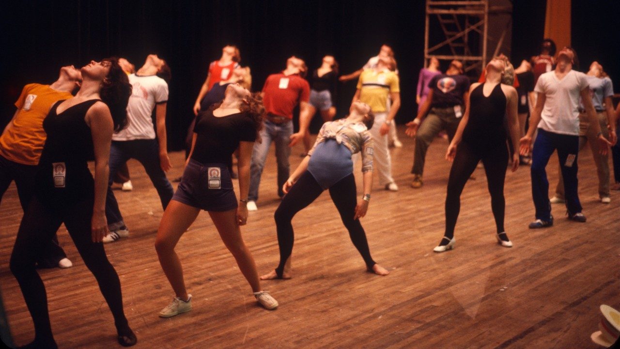  A group of New Virginians warms up on stage. They all wear various warmup or exercise clothes and a nametag pinned to their clothing. They all lean their heads back, front leg bent slightly and back leg supporting their weight, arms at their sides.
