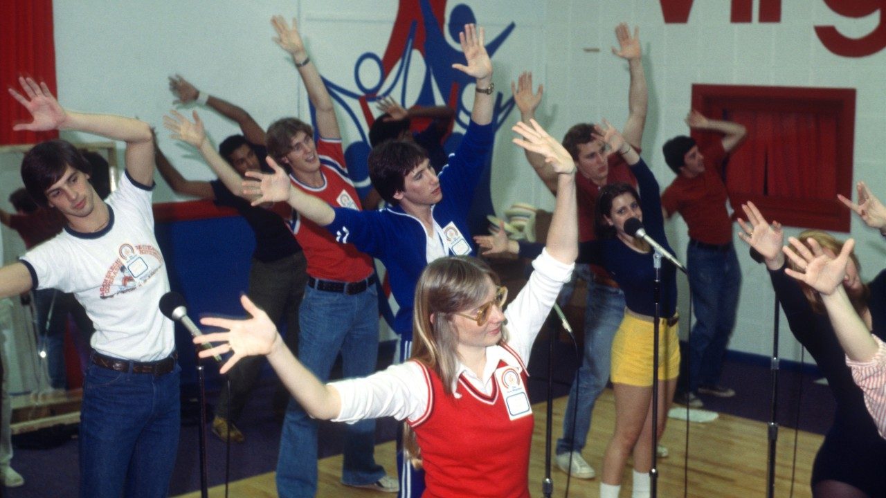  Members of the New Virginians rehearse or perform in a room with a hardwood floor and white walls with the New Virginians logo painted on it in red and blue. Everyone stands behind microphone stands and lifts their arms above their heads, stretched to their right sides.