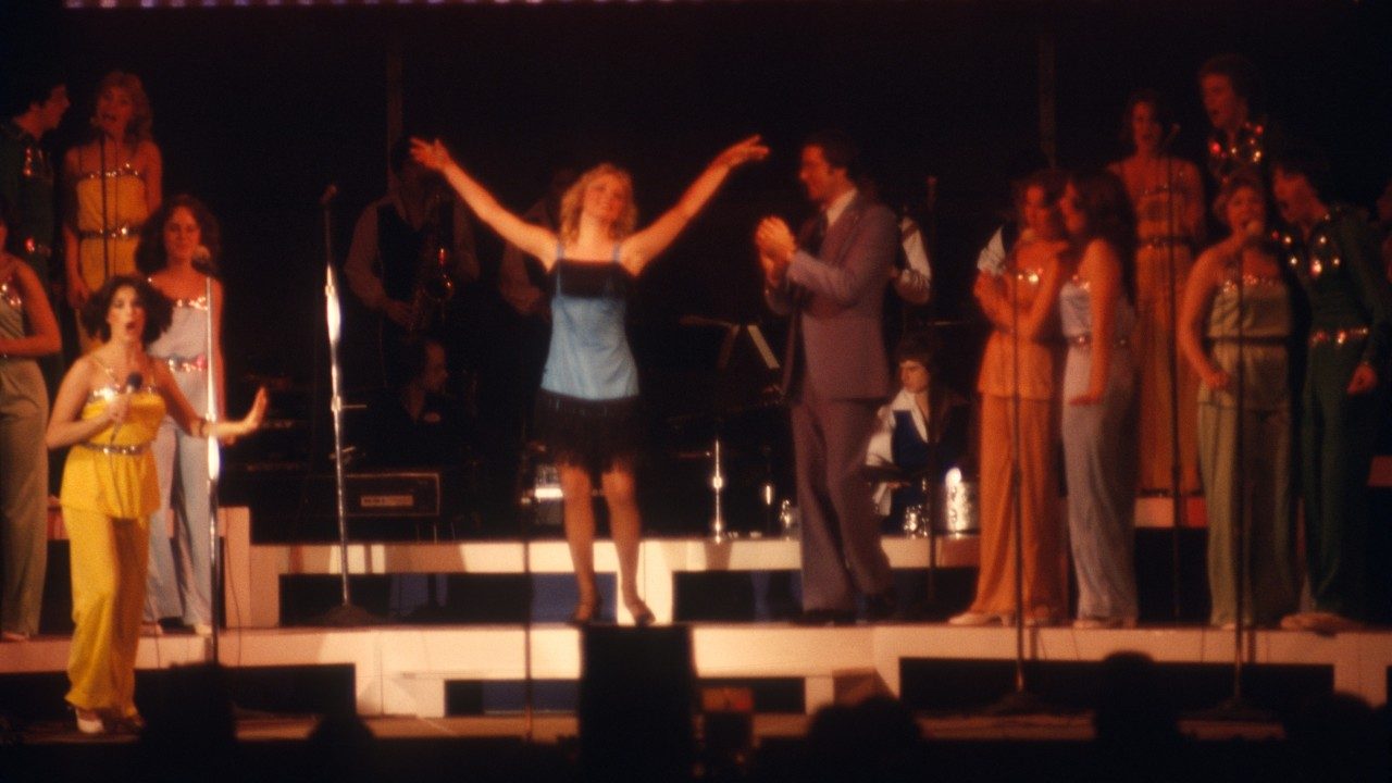  Members of the New Virginians perform on stage in this somewhat blurry photo. In the center of the frame, a white woman with medium length blonde hair wears a blue flapper-inspired dress with black fringe at the top and bottom, and raises both of her arms elegantly into the air. The other women on stage wear jumpsuits in various colors with sequins at their waists and necklines.