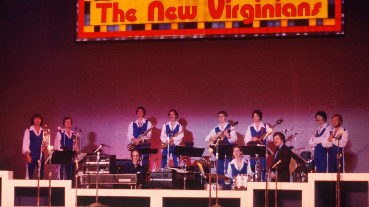  Members of the New Virginians hold their instruments on stage in front of a maroon or purple wall. Above them is a light up "The New Virginians" sign. Almost all of them wear blue jumpsuits over white button down shirts.