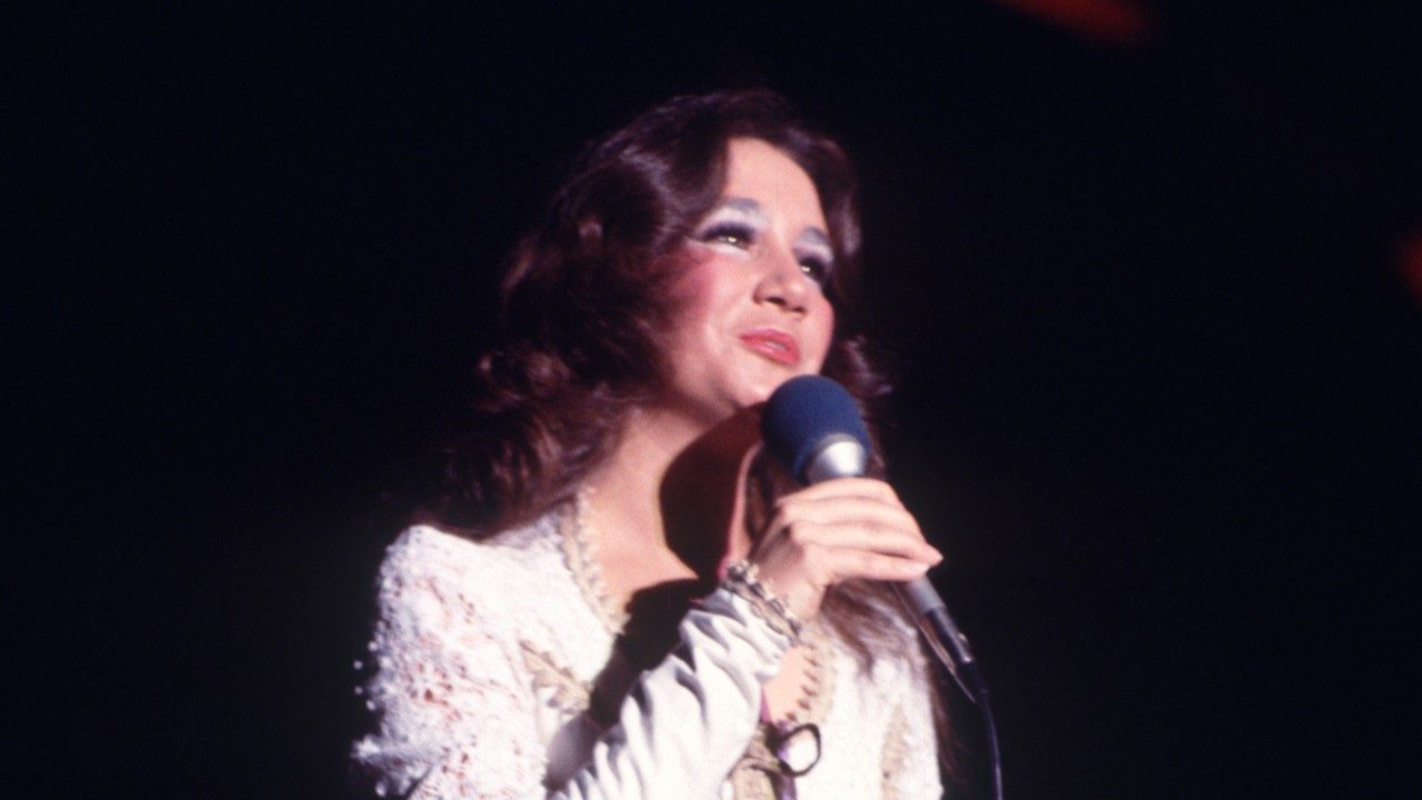  A member of the New Virginians, a white woman with long dark brown hair, sings into a microphone wearing a white longsleeved dress. Her eyeshadow is white and she wears a berry-colored lipstick.