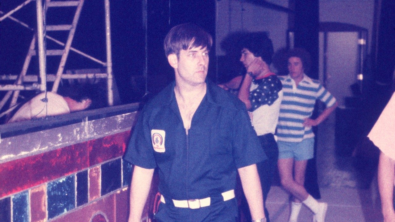  Members of the New Virginians backstage. In the center and foreground is a white man with dark brown hair. He wears a navy blue pair of coveralls with a white belt and the New Virginians logo on the right breast pocket.