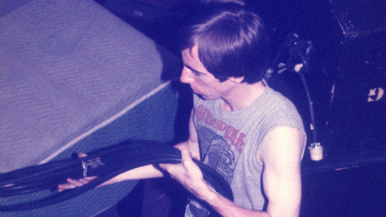  A member of the New Virginians wrangles a bundle of cables. He is a white man with medium length dark brown hair. He wears a sleeveless grey t-shirt.