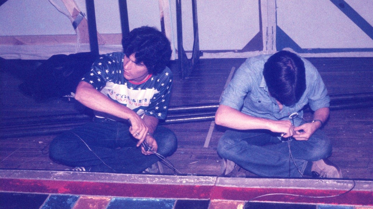  Two members of the New Virginians' tech crew sit cross-legged on the floor while they work on the wiring for a light up sign. Both are white men with dark brown hair. The one of the left wears a navy blue shirt with white stars on it and jeans. The one of the right wears a grey-blue button down shirt with jeans.