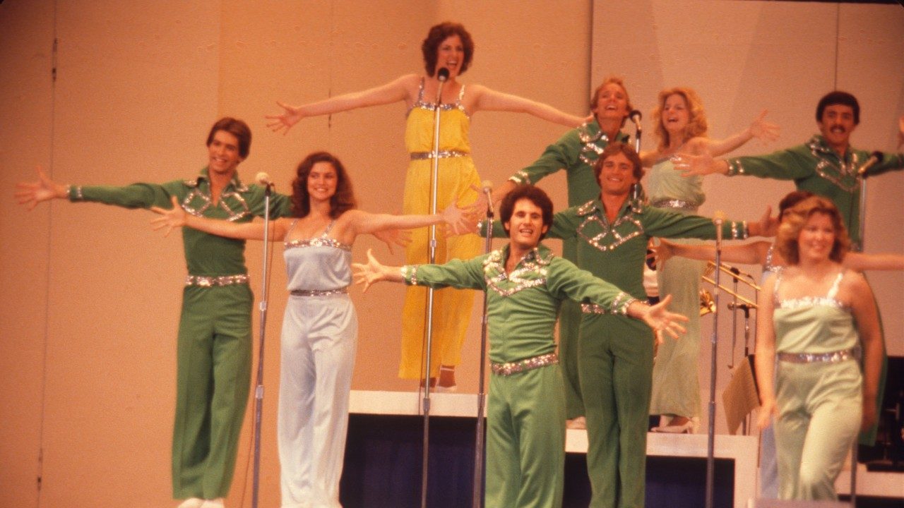  Members of the New Virginians perform on stage. They sing into microphones, almost everyone with both arms raised and extended from their bodies in a T shape. The men wear kelly green jumpsuits with sequined belts and geometric details on the chest. The women wear jumpsuits of various colors (light purple, mustard, sage green), with sequins at the belt and the top trim.