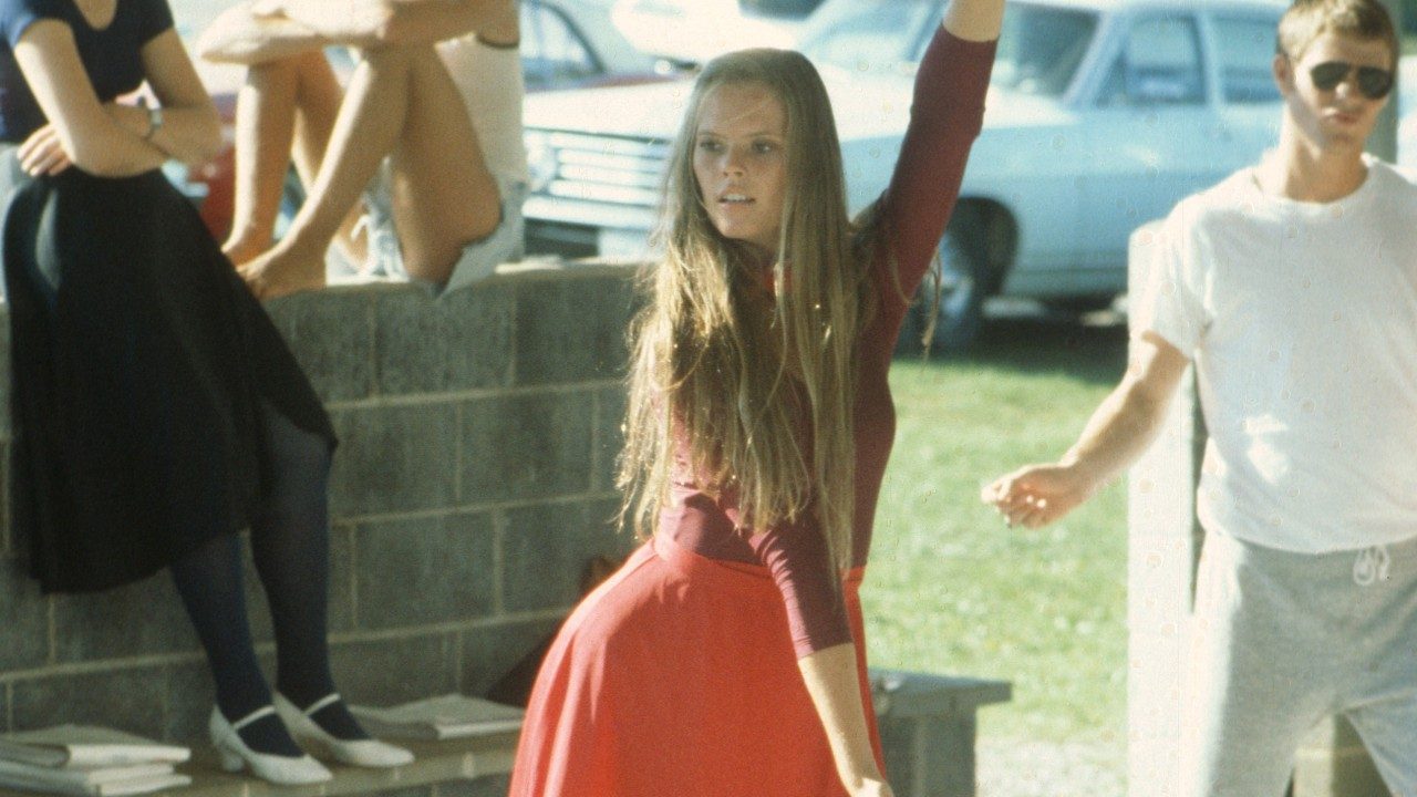  A member of the New Virginians performs in an outdoor area. She is a white woman with long light brown hair wearing a maroon longsleeved shirt (the sleeves are pushed up her forearms) and an orange A-line skirt. She looks to her right with her hips jutted to the right, right arm straight and pointed down in front of her and left arm raised.
