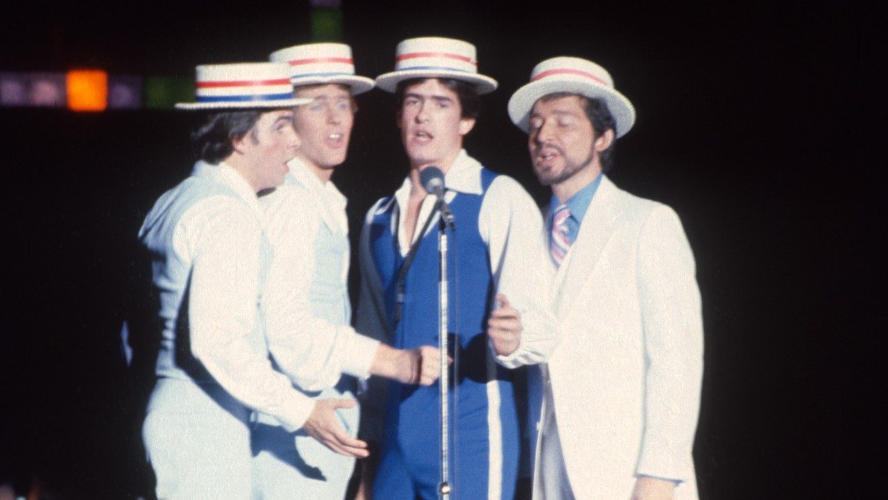  A group of four New Virginians, white men dressed like a barbershop quartet, sing into a microphone on a stand.