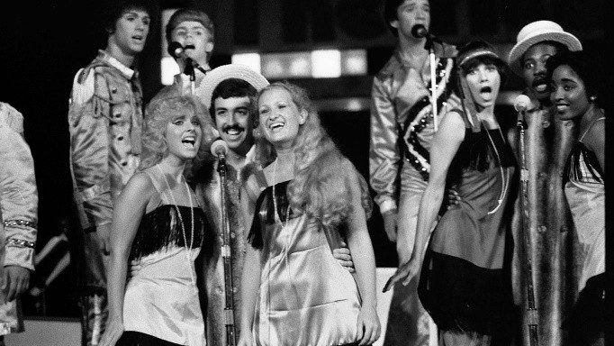  Members of the New Virginians perform on stage. In the foreground, two sets of trios sing into the microphones, the women wearing flapper costumes, and the men wearing white top hats.