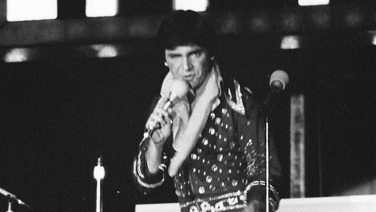  A member of the New Virginians, a white man with dark brown hair wearing a dark and sequined Elvis costume, performs on stage. He is blurred a bit from his movement.