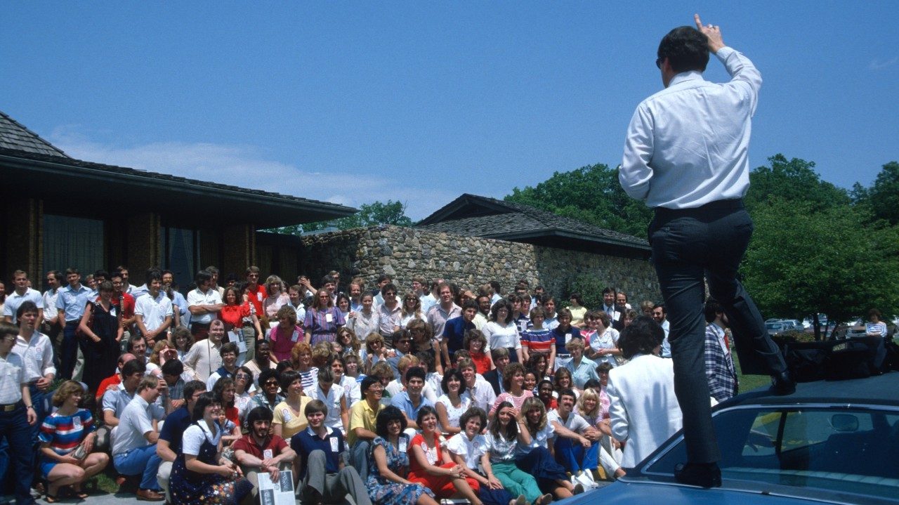  In the foreground, a white man with dark brown hair, a light button down shirt, and dark pants stands on top of a car, right hand raised above his head. He's facing a large group of New Virginians, assembled in front of a low-slung stone building, all sitting, crouching, or standing for a group photo.