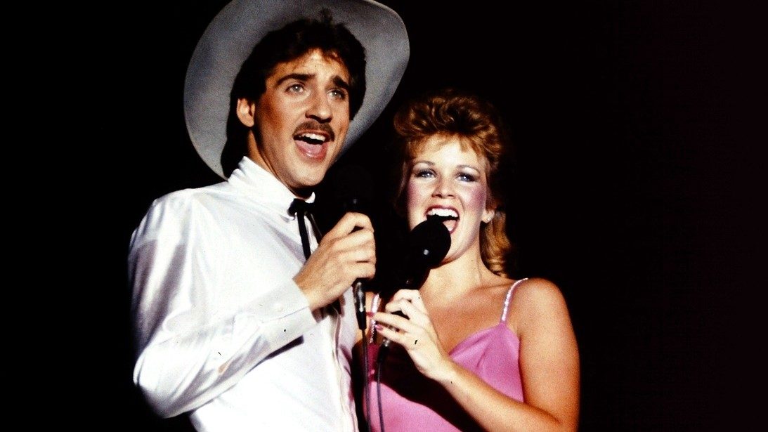  Two members of the New Virginians perform on stage in this color image. On the left is a white man with medium length dark brown hair and a brown moustache. He wears a white button down shirt, black bolo tie, and large white cowboy hat. On the right is a white woman with medium length red hair wearing a magenta spaghetti strap dress.