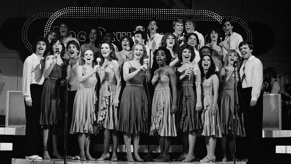  The New Virginians perform as a group on stage in this black and white image from 1982. The women wear knee-length dresses with ruffles and the men wear white shirts with dark pants. They stand on stage risers.