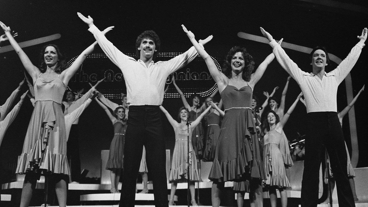  Members of the New Virginians perform on stage in this black and white photo. In the foreground are four people, two white women and two white men in alternating order. They stand with their legs in a slightly wide stance and arms thrown wide over their heads in an X shape. Behind them are more members on stage risers in the same stance. The men wear white shirts tucked into dark pants; the women wear A-line dresses with spaghetti straps, sweetheart necklines, thin belts at the waist, and a line of ruffles cascading from the belt to the ruffled hem.