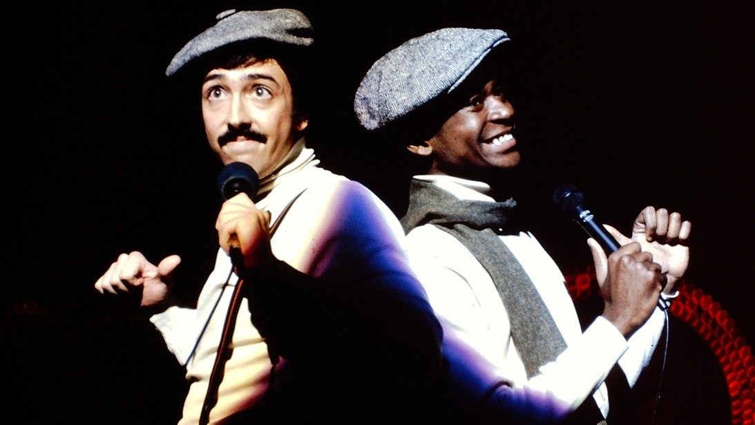  Two members of the New Virginians perform on stage. On the left is a white man with dark hair and a dark moustache wearing a white turtleneck and grey hat; on the left is a Black man wearing a white shirt, grey scarf, and grey hat. Both point their thumbs at themselves. They are lit by by individual follow spots and lean their backs against each other.