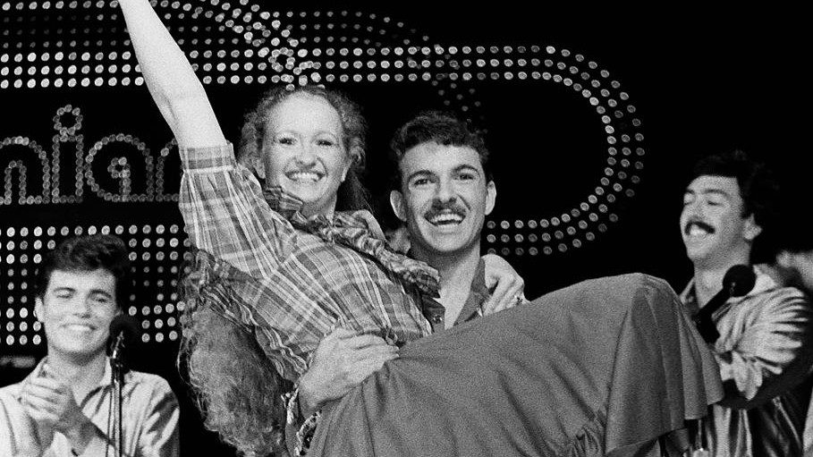  Members of the New Virginians perform on stage in this black and white photo. In the foreground, a white man holds a white woman in his arms. She wears a plaid shirt and long ruffled skirt, almost like a woman on the American frontier would wear, and raises her right arm above her head. Behind them are other members of the group and part of the New Virginians' light-up sign.