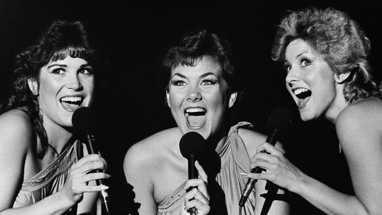  Three members of the New Virginians perform on stage in this black and white photo. All three are white woman, two with dark brown hair and one with blonde hair.  They wear satin dresses tied at one shoulder and sing animatedly.