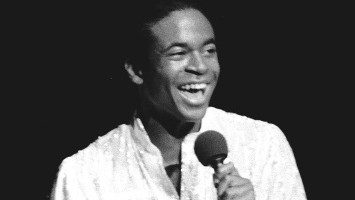 A member of the New Virginians, a Black man wearing an oversized white sequined button down shirt, wide silver belt, and dark pants, sings into a microphone and snaps his fingers in this black and white photo.