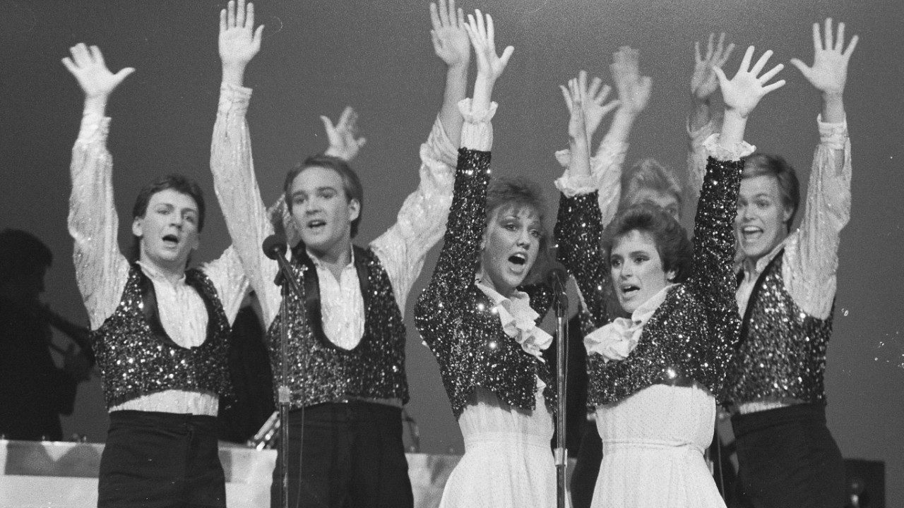  Six members of the New Virginians, four white men and two white women, perform and sing on stage in this black and white image. The men wear white button down shirts under sequined vests with black pants. The women wear white shirts and long skirts under sequined jackets. All of them raise their hands into the air.