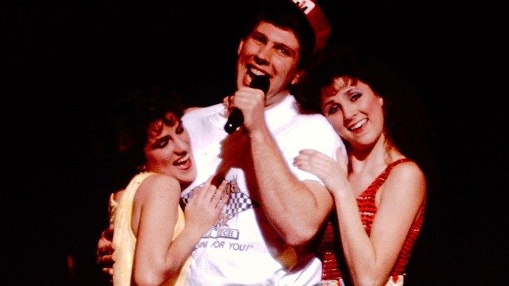  Three members of the New Virginians perform on stage. In the middle is a tall white man with dark brown hair, maroon baseball cap, white shirt, brown belt, and dark jeans. Two white women with dark brown hair hug him from either side, one leg kicked up at a 90 degree angle flirtatiously.