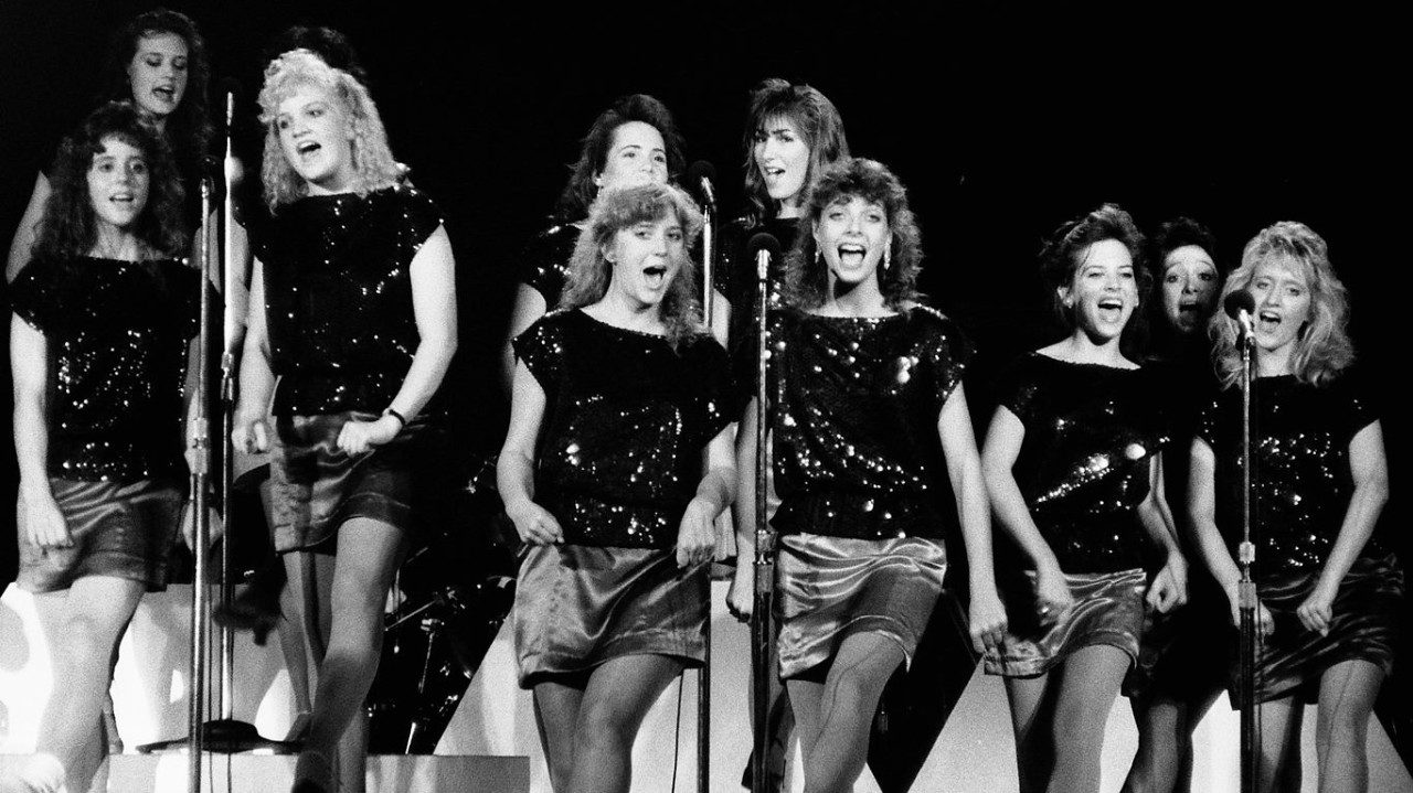  Members of the New Virginians wear sequined black tops and satin skirts while singing by pairs into several microphones in this black and white photo.