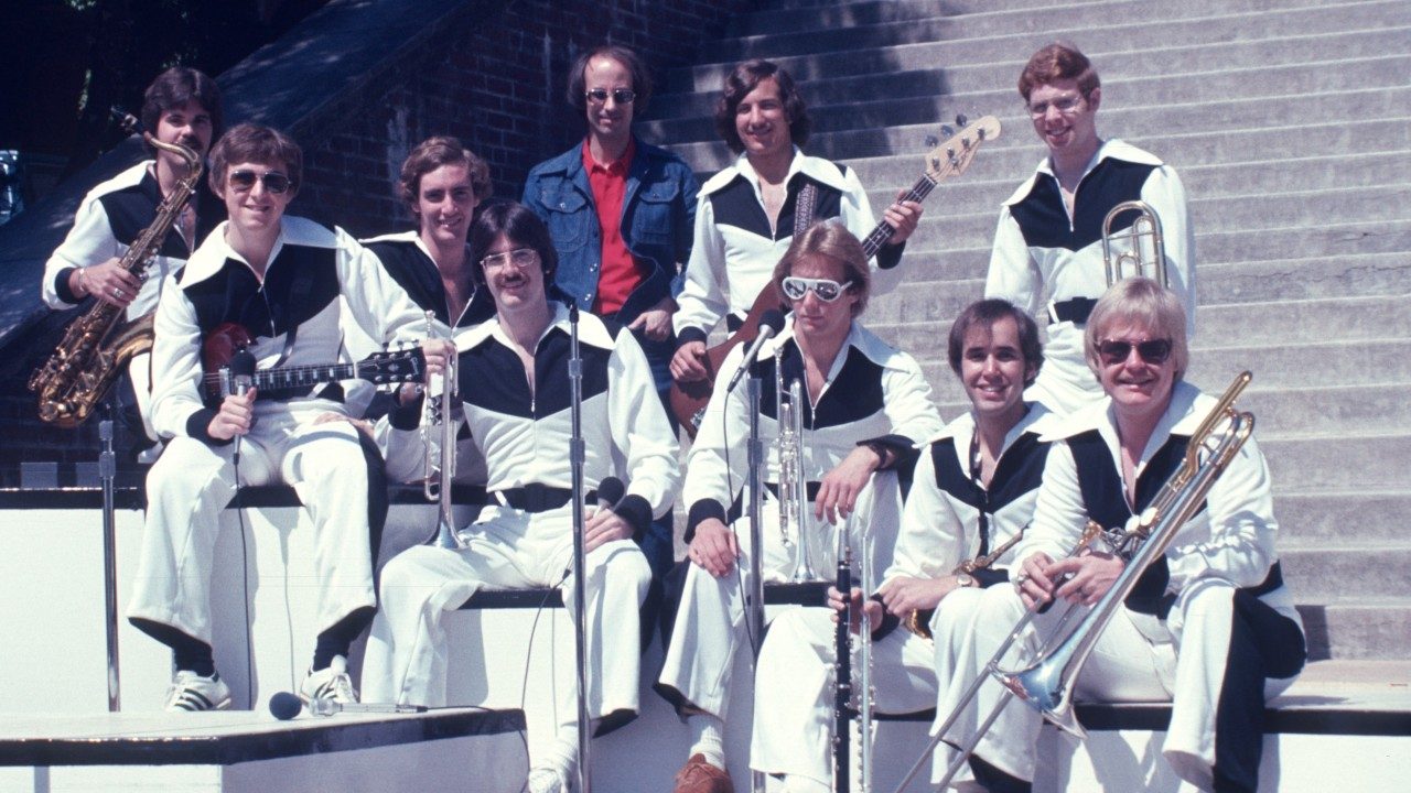  A group of male New Virginians hold their instruments and sit on a set piece on the landing of a set of concrete stairs outside. They all wear white pants with a chunky black stripe running down the outside of the leg, and white shirts with really wide lapels, black detail on the chest, and black belts. In the center in the back is a mean in a jean jacket and red shirt.